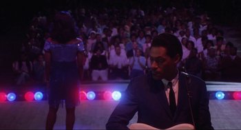 Movie still from “What's Love Got to Do with It” (1993), directed by Brian Gibson – A man sitting in front of an audience in front of a crowd; Medium shot, Low angle
