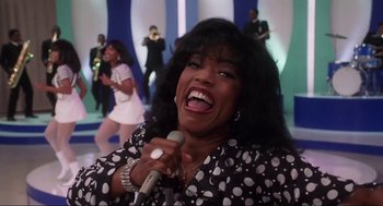 Movie still from “What's Love Got to Do with It” (1993), directed by Brian Gibson – A woman is singing with a microphone in front of a band; Close Up shot, Low angle