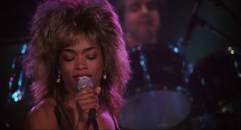 Movie still from “What's Love Got to Do with It” (1993), directed by Brian Gibson – A woman with a big hair wig is holding a microphone in front of a band; Close Up shot, Over the shoulder angle
