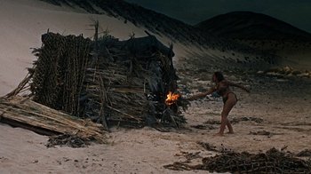Movie still from “When Dinosaurs Ruled the Earth” (1970), directed by Val Guest – A woman in a bathing suit standing next to a pile of wood; Wide shot, High angle