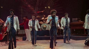 Movie still from “When We Were Kings” (1996), directed by Leon Gast – A group of men singing on a stage with a microphone; Wide shot, High angle