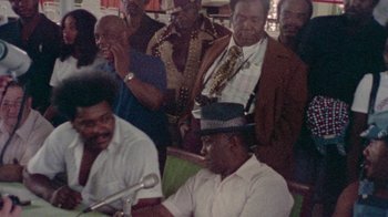 Movie still from “When We Were Kings” (1996), directed by Leon Gast – A group of men standing next to each other in a room; Medium shot, High angle