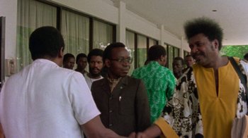 Movie still from “When We Were Kings” (1996), directed by Leon Gast – A group of people standing around each other in front of a building; Medium shot, Over the shoulder angle