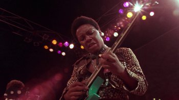Movie still from “When We Were Kings” (1996), directed by Leon Gast – A man holding a green guitar in front of a crowd; Medium shot, Low angle