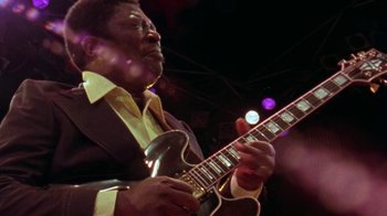 Movie still from “When We Were Kings” (1996), directed by Leon Gast – A man in a suit playing an electric guitar on a stage; Close Up shot, Low angle
