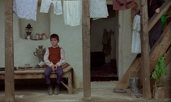 Movie still from “Where Is the Friend's House?” (1987), directed by Abbas Kiarostami – A young boy sitting on a wooden bench in a room; Wide shot, Over the shoulder angle