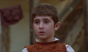 Movie still from “Where Is the Friend's House?” (1987), directed by Abbas Kiarostami – A young boy wearing a brown vest looks at the camera; Close Up shot, Over the shoulder angle