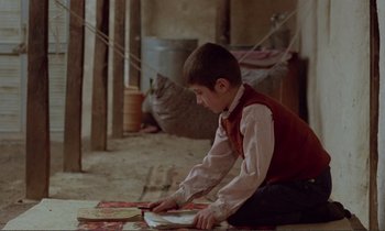 Movie still from “Where Is the Friend's House?” (1987), directed by Abbas Kiarostami – A young boy sitting on the ground with a book; Wide shot, High angle