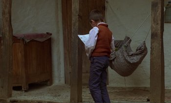 Movie still from “Where Is the Friend's House?” (1987), directed by Abbas Kiarostami – A young boy in a red vest and blue pants holding a paper; Wide shot, High angle