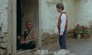 Movie still from “Where Is the Friend's House?” (1987), directed by Abbas Kiarostami – An old man sitting next to a young boy in front of a building; Wide shot, Over the shoulder angle