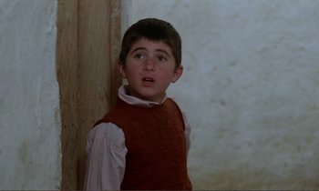 Movie still from “Where Is the Friend's House?” (1987), directed by Abbas Kiarostami – A young boy wearing a red vest standing next to a wall; Close Up shot, Over the shoulder angle
