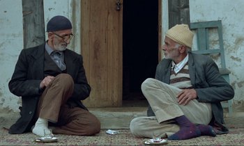Movie still from “Where Is the Friend's House?” (1987), directed by Abbas Kiarostami – Two older men sitting on the ground in front of a building; Wide shot, High angle