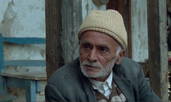 Movie still from “Where Is the Friend's House?” (1987), directed by Abbas Kiarostami – An older man wearing a hat sitting in front of a wooden pole; Close Up shot, Over the shoulder angle