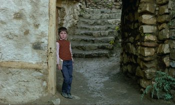 Movie still from “Where Is the Friend's House?” (1987), directed by Abbas Kiarostami – A young boy standing in a doorway of a stone house; Wide shot, Low angle