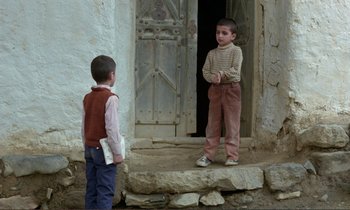 Movie still from “Where Is the Friend's House?” (1987), directed by Abbas Kiarostami – Two young boys standing in front of an old building; Wide shot, Over the shoulder angle