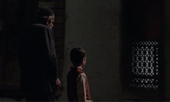 Movie still from “Where Is the Friend's House?” (1987), directed by Abbas Kiarostami – A man standing next to a young boy in front of a brick wall; Medium shot, Over the shoulder angle