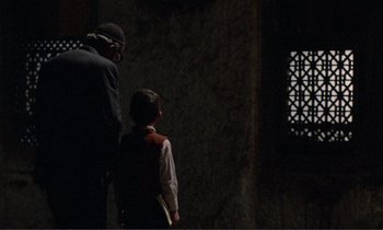 Movie still from “Where Is the Friend's House?” (1987), directed by Abbas Kiarostami – A man standing next to a child in a dark room; Medium shot, Over the shoulder angle