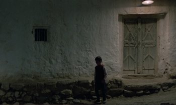 Movie still from “Where Is the Friend's House?” (1987), directed by Abbas Kiarostami – A young boy standing in front of a building; Wide shot, High angle