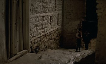 Movie still from “Where Is the Friend's House?” (1987), directed by Abbas Kiarostami – A person standing in a dark alley near a stone wall; Extreme Wide shot, High angle