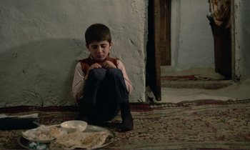 Movie still from “Where Is the Friend's House?” (1987), directed by Abbas Kiarostami – A boy sitting on the floor with a plate of food in front of him; Medium shot, High angle