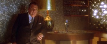 Movie still from “Where the Truth Lies” (2005), directed by Atom Egoyan – A man sitting at a table in a room with a lamp; Medium shot, Low angle