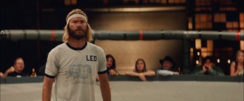 Movie still from “Whip It” (2009), directed by Drew Barrymore – A man with long blonde hair standing in front of a group of people; Medium shot, Low angle