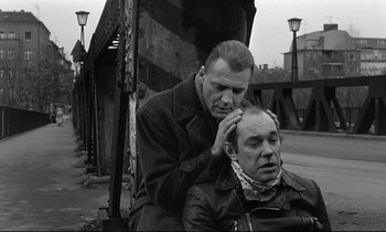Movie still from “Wings of Desire” (1987), directed by Wim Wenders – A man sitting next to another man on the side of a road; Medium shot, Over the shoulder angle