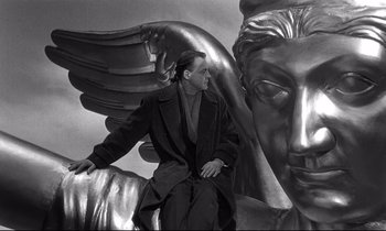 Movie still from “Wings of Desire” (1987), directed by Wim Wenders – A black and white photo of a man sitting in front of a statue of an angel; Medium shot, Low angle