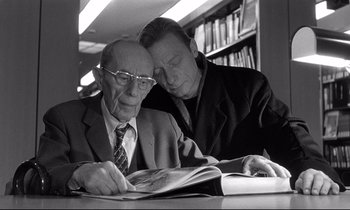 Movie still from “Wings of Desire” (1987), directed by Wim Wenders – A man reading a magazine with another man looking on; Close Up shot, Low angle