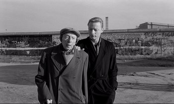 Movie still from “Wings of Desire” (1987), directed by Wim Wenders – A man standing next to an older man in front of a wall; Medium shot, Over the shoulder angle