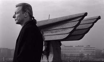 Movie still from “Wings of Desire” (1987), directed by Wim Wenders – A man standing next to a bench on a cloudy day; Medium shot, Low angle
