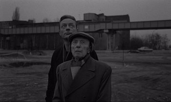 Movie still from “Wings of Desire” (1987), directed by Wim Wenders – A man standing next to an older woman in front of a building; Medium shot, Low angle