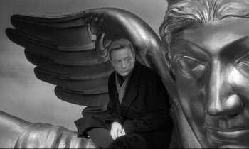 Movie still from “Wings of Desire” (1987), directed by Wim Wenders – A black and white photo of a man sitting on a wing of an angel; Medium shot, High angle