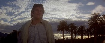 Movie still from “Winter Kills” (1979), directed by William Richert – A man standing in front of palm trees under a cloudy sky; Close Up shot, Low angle