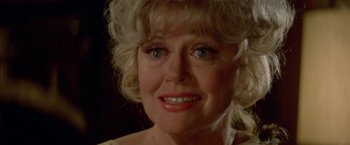 Movie still from “Winter Kills” (1979), directed by William Richert – An older woman with blonde curly hair wearing a necklace; Close Up shot, Over the shoulder angle