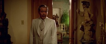 Movie still from “Winter Kills” (1979), directed by William Richert – An older man in a white suit standing in front of a doorway; Medium shot, Over the shoulder angle