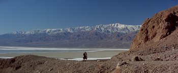 Movie still from “Winter Kills” (1979), directed by William Richert – Two people standing on a beach near a body of water and mountains; Extreme Wide shot, Low angle