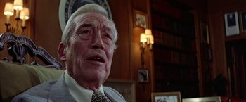 Movie still from “Winter Kills” (1979), directed by William Richert – An older man in a suit and tie in front of a bookshelf; Close Up shot, Over the shoulder angle