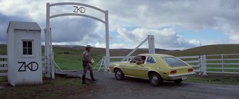 Movie still from “Winter Kills” (1979), directed by William Richert – A man walking next to a yellow car on a dirt road; Wide shot, Over the shoulder angle
