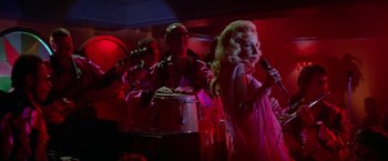 Movie still from “Winter Kills” (1979), directed by William Richert – A group of people in a room singing and playing musical instruments; Medium shot, Over the shoulder angle
