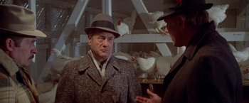 Movie still from “Winter Kills” (1979), directed by William Richert – A man wearing a hat talking to another man in front of chickens; Medium shot, Over the shoulder angle