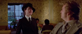 Movie still from “Winter Kills” (1979), directed by William Richert – A woman wearing a suit and a hat in front of other people; Medium shot, Over the shoulder angle