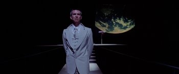 Movie still from “Winter Kills” (1979), directed by William Richert – A man in a white suit standing in front of an image of the earth; Medium shot, Low angle