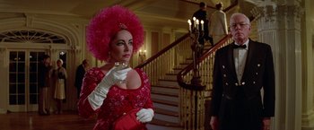 Movie still from “Winter Kills” (1979), directed by William Richert – A woman in a red dress holding a wine glass in front of a staircase; Medium shot, Over the shoulder angle
