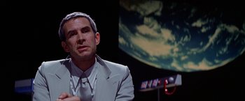 Movie still from “Winter Kills” (1979), directed by William Richert – A man wearing a suit and tie in front of a screen; Close Up shot, Over the shoulder angle