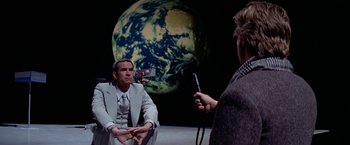 Movie still from “Winter Kills” (1979), directed by William Richert – A man is being interviewed in front of a globe; Medium shot, Over the shoulder angle