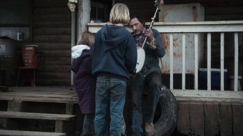 Movie still from “Winter's Bone” (2010), directed by Debra Granik – A man playing a banjo on a porch with two children; Medium shot, Over the shoulder angle