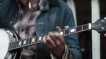 Movie still from “Winter's Bone” (2010), directed by Debra Granik – A man is holding a guitar and smoking a cigarette; Extreme Close Up shot, Over the shoulder angle