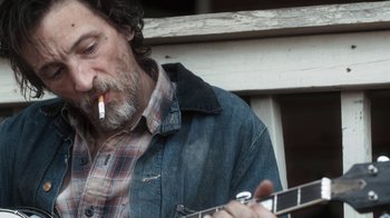 Movie still from “Winter's Bone” (2010), directed by Debra Granik – A man smoking a cigarette while holding a guitar; Close Up shot, Over the shoulder angle