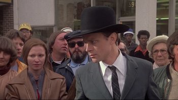Movie still from “Wise Blood” (1979), directed by John Huston – A man wearing a hat and a suit walking down a street; Close Up shot, Over the shoulder angle